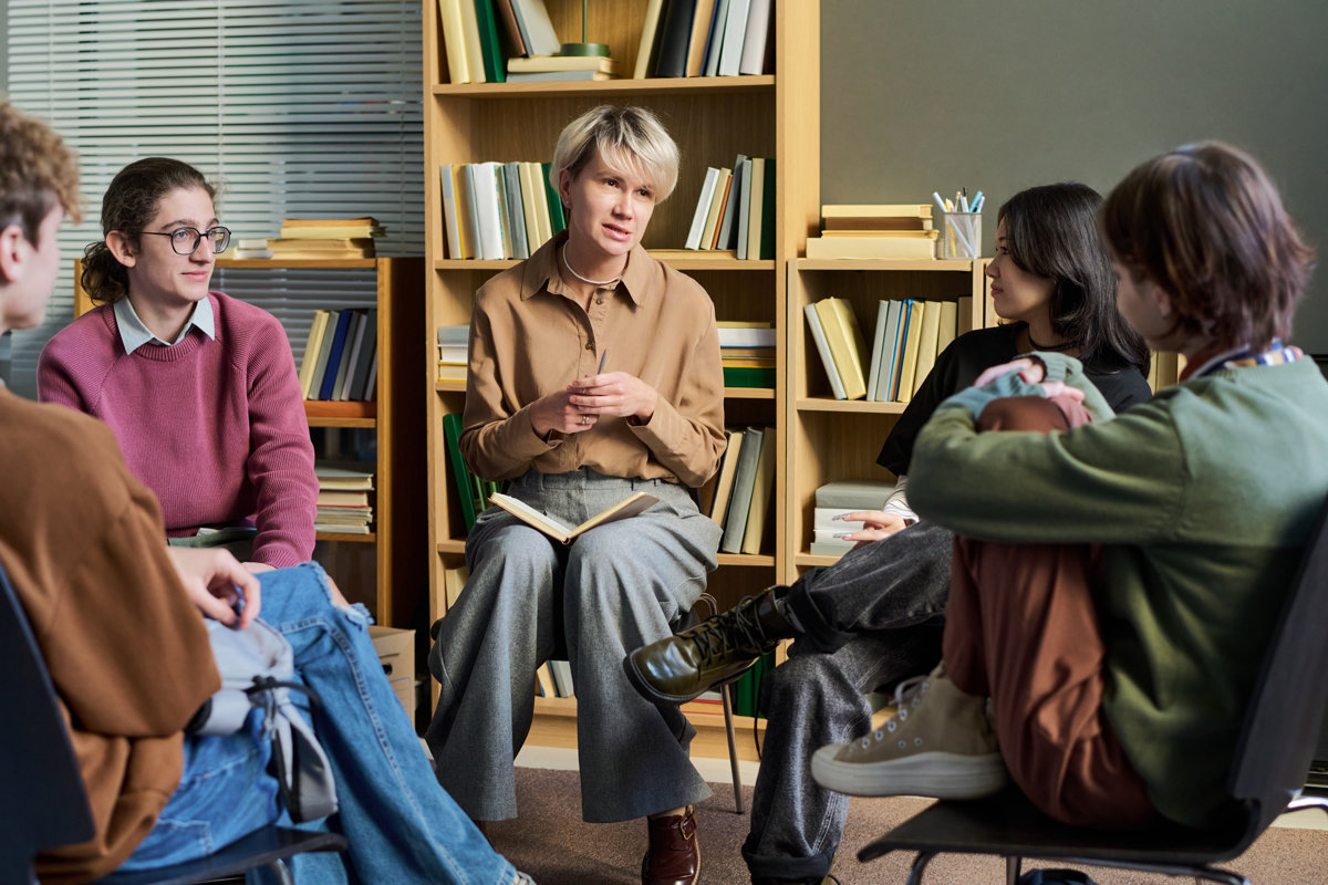 Group of diverse teenagers with adult woman sitting in circle discussing during group therapy session, participants in conversation and listening attentively