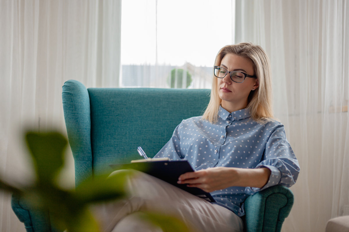 Woman psychologist or psychotherapist with eyeglasses sitting in armchair and writes on clipboard