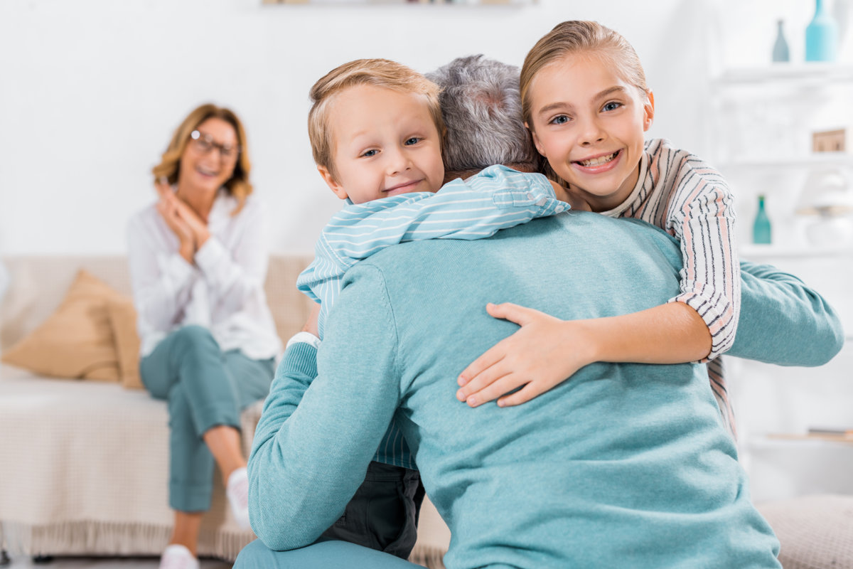 selective focus of smiling kids looking at camera and hugging grandfather at home